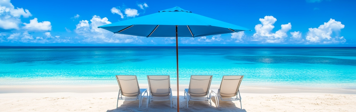 Four lounge chairs under a large blue umbrella on a pristine white sandy beach, overlooking the clear waters of the ocean.